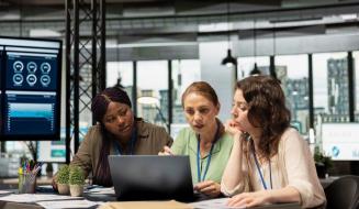 Três mulheres de diferentes etnias em uma reunião de trabalho, sentadas ao redor de uma mesa com um laptop, papéis e pequenos vasos de plantas.