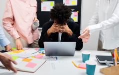 Profissional com a cabeça entre as mãos demonstra estresse enquanto colegas ao redor pressionam com demandas, sugerindo sobrecarga ou assédio no trabalho.