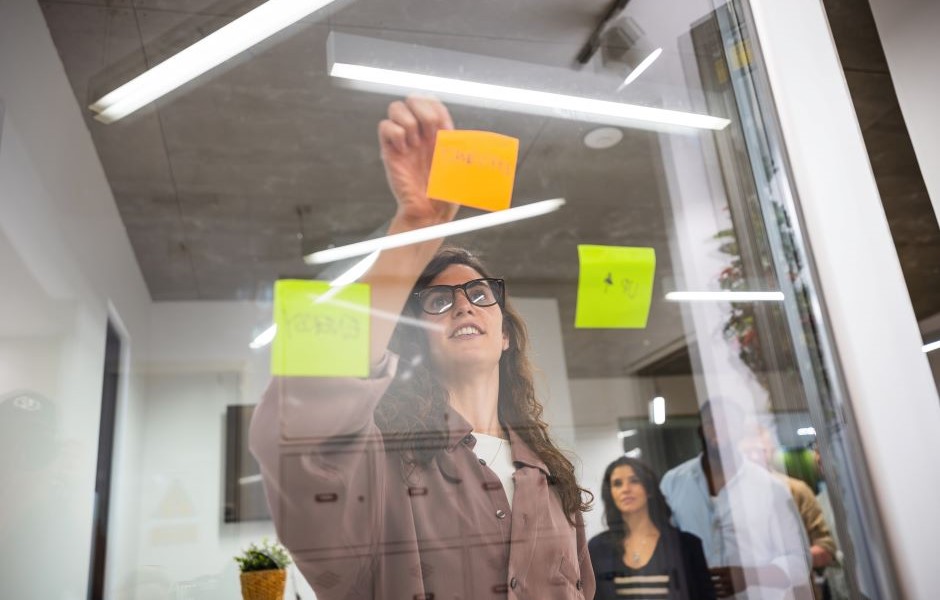 Mulher de óculos escrevendo lembretes em post-its coloridos na parede de vidro em ambiente empresarial, promovendo organização e produtividade