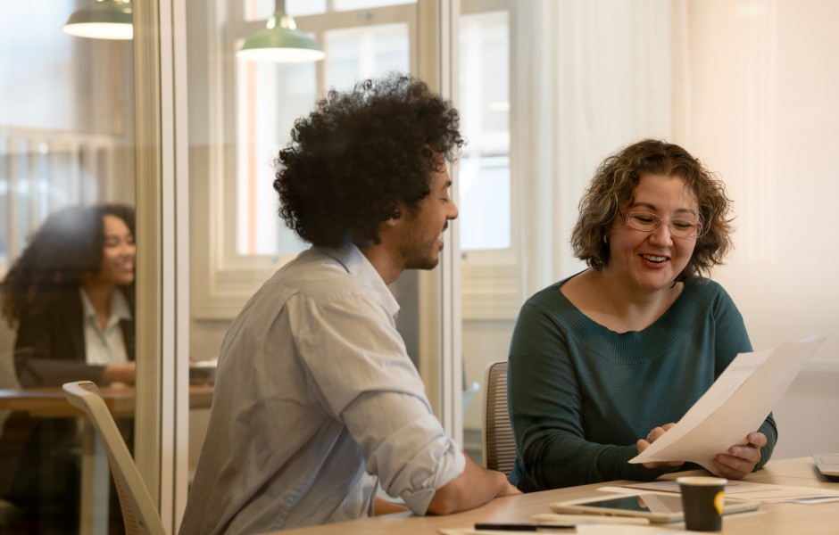 Dois colegas de trabalho, um homem com cabelo afro e uma mulher de óculos, conversam de forma animada e sorridente enquanto analisam um documento impresso em uma sala de escritório com divisórias de vidro.