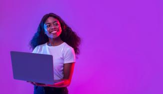 ma jovem negra sorridente segura um notebook aberto, iluminada por luzes vibrantes em tons de roxo e rosa neon.