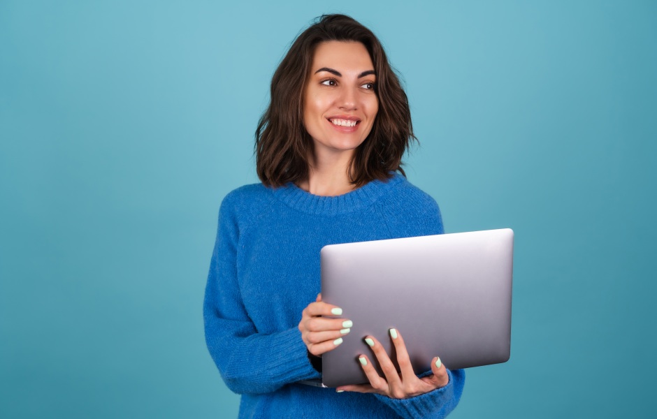 Uma jovem de blusa de tricô azul segura um notebook e olha para o lado com um sorriso alegre, sobre fundo azul.