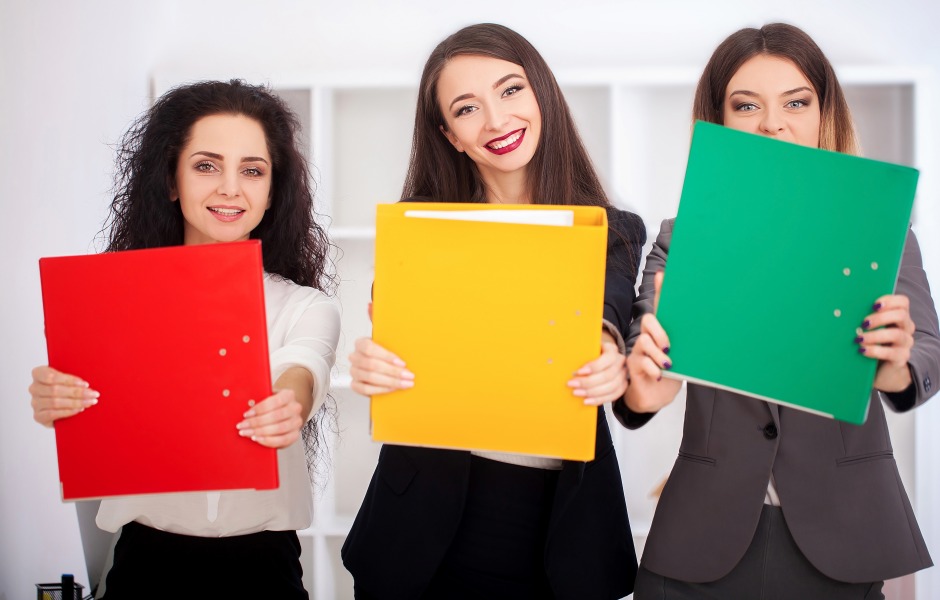 Três mulheres sorridentes seguram pastas coloridas (vermelha, amarela e verde) à frente do corpo em um ambiente de escritório.
