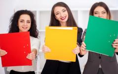 Três mulheres sorridentes seguram pastas coloridas (vermelha, amarela e verde) à frente do corpo em um ambiente de escritório.