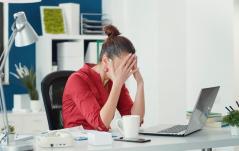 Mulher sentada em uma mesa de escritório com as mãos cobrindo o rosto, demonstrando cansaço ou estresse diante de um notebook aberto e uma xícara de café.