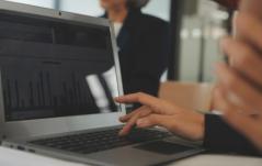 Mãos digitando em notebook com gráfico de barras na tela, em ambiente de reunião corporativa.