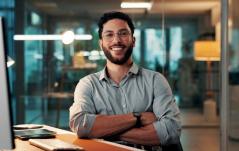 Homem jovem com óculos e barba, sorrindo com os braços cruzados, sentado em uma mesa de escritório moderno com iluminação noturna ao fundo.