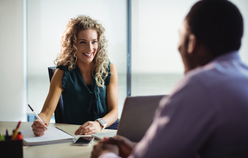 Mulher sorridente anota em caderno enquanto conversa com um colega, sugerindo uma reunião one-on-one ou entrevista profissional.