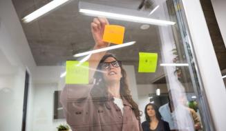 Mulher de óculos escrevendo lembretes em post-its coloridos na parede de vidro em ambiente empresarial, promovendo organização e produtividade