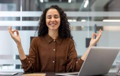 Mulher meditando com olhos fechados e sorrindo à mesa do escritório, transmitindo tranquilidade e equilíbrio no trabalho.