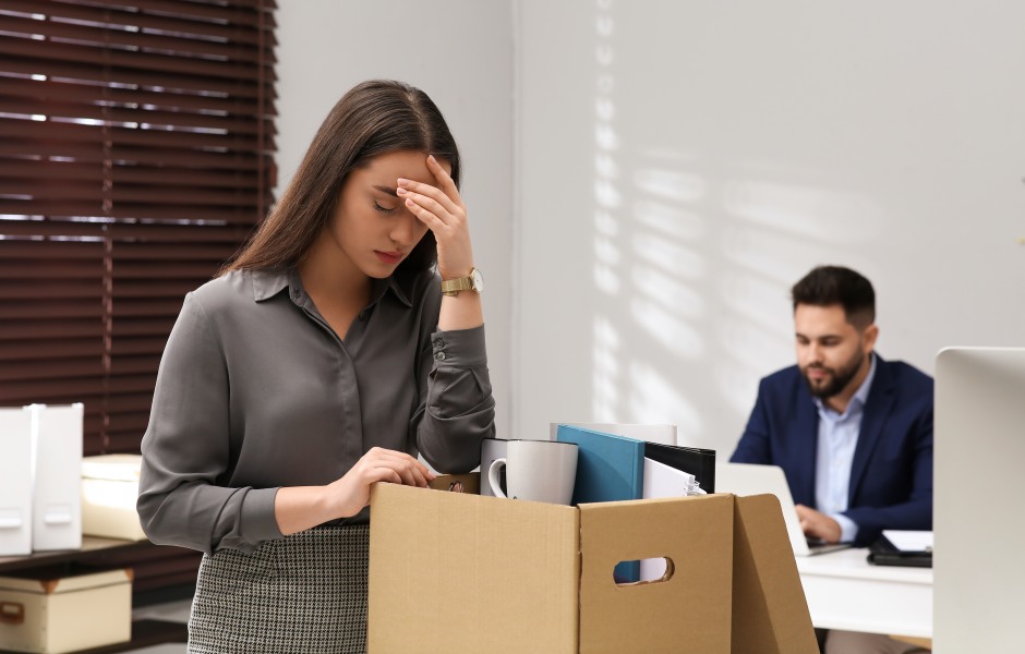 Mulher abatida recolhendo seus pertences em uma caixa no escritório, representando uma demissão.