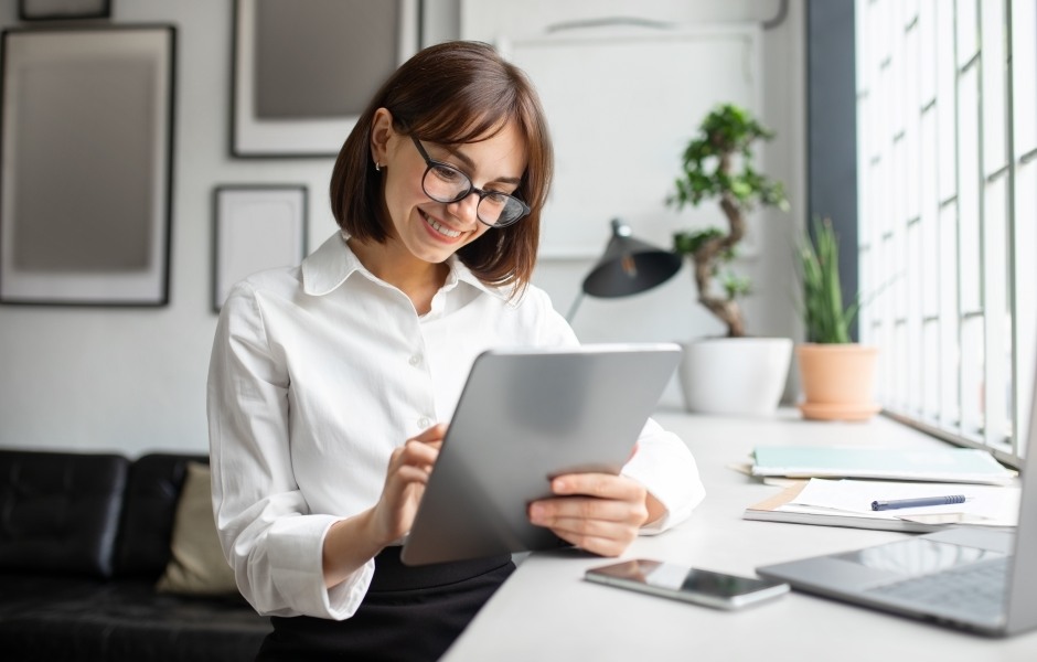 Mulher de óculos e camisa branca sorri ao usar um tablet em um escritório moderno e bem iluminado.