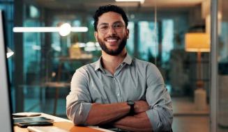 Homem jovem com óculos e barba, sorrindo com os braços cruzados, sentado em uma mesa de escritório moderno com iluminação noturna ao fundo.