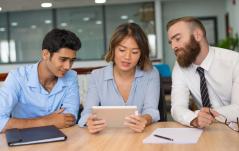 Três colegas analisam juntos um tablet em uma mesa, em ambiente de escritório colaborativo.