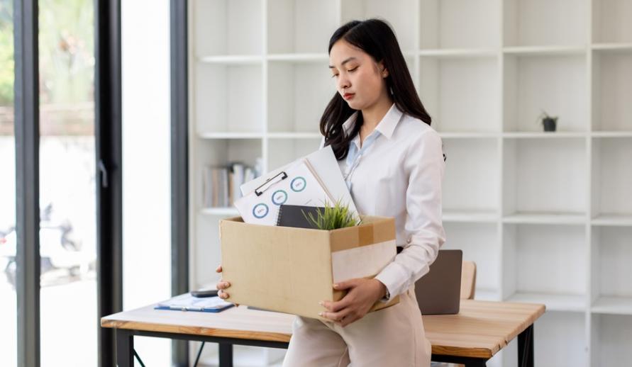 Mulher vestindo camisa social branca, segurando uma caixa com documentos e uma planta, em ambiente de escritório moderno. A imagem representa a organização no trabalho.